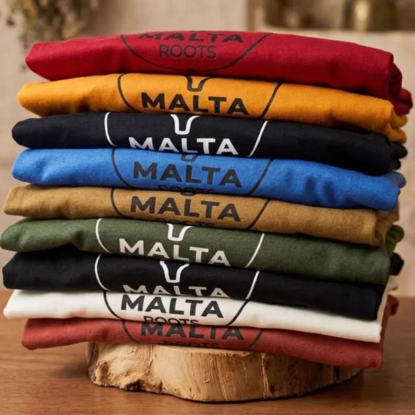Camisetas Malta Roots empilhadas em várias cores sobre tronco de madeira